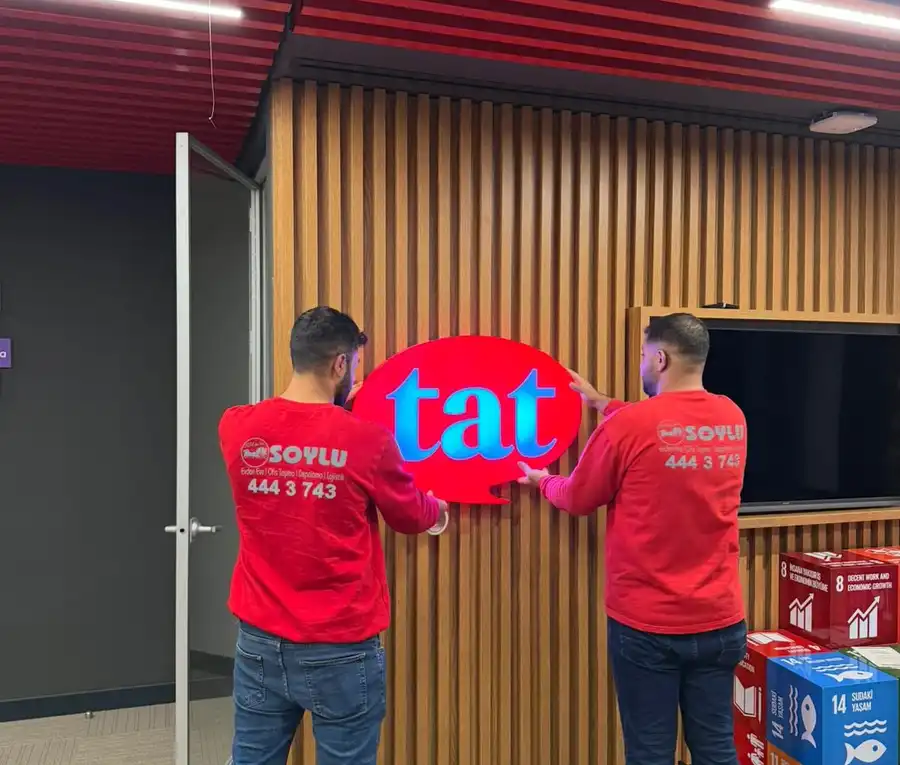 Kırmızı üniformalı Soylu Nakliyat ekibi, taşınan şirketin logo tabelasını yeni ofisin duvarına yerleştiriyor
