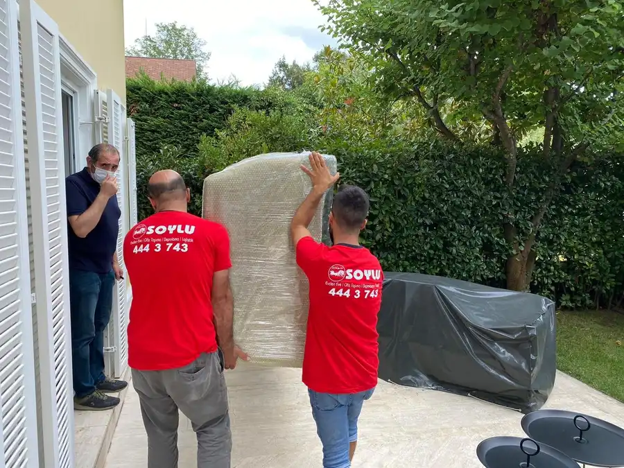 İki Soylu çalışanı balonlu naylona sarılı bir eşyayı bahçe terasında taşıyor, kapıda bir kişi izliyor