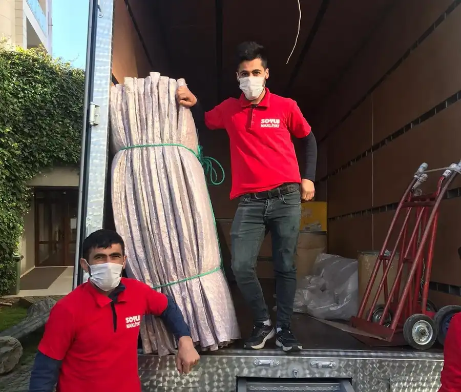 Kamyon kasasında iple bağlanmış taşıma battaniyesi demeti ve el arabalarının yanında duran maskeli iki taşıma personeli
