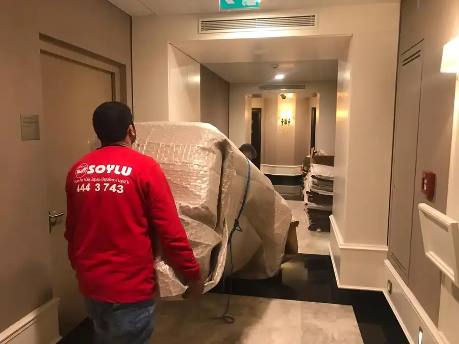 Bina koridorunda balonlu naylona sarılı büyük bir eşyayı taşıyan iki Soylu Nakliyat personeli