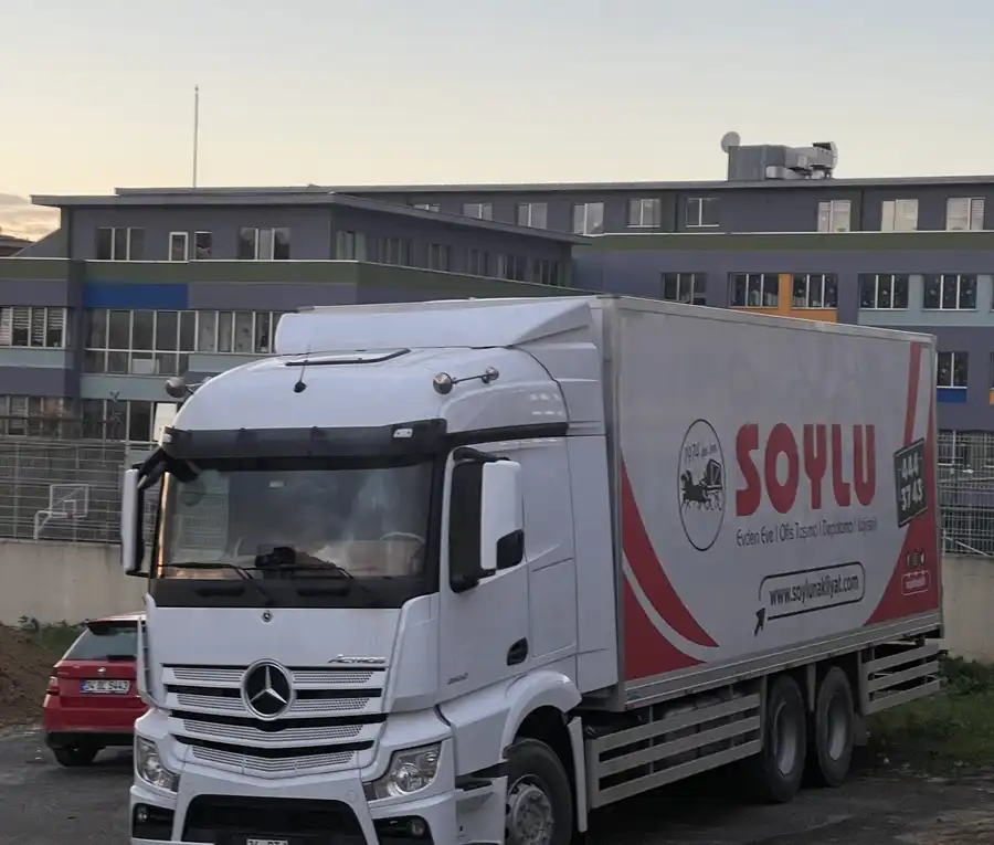 Kapalı kasalı büyük beyaz Soylu kamyonu akşamüstü toprak bir alanda park etmiş, arkada renkli cepheli bir bina