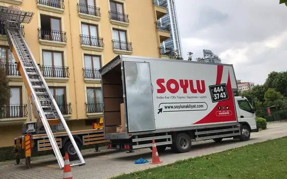 Ferforje balkonlu sarı bir apartmanın üst katlarına uzanan eşya asansörü, yanında kasası açık Soylu kamyonu
