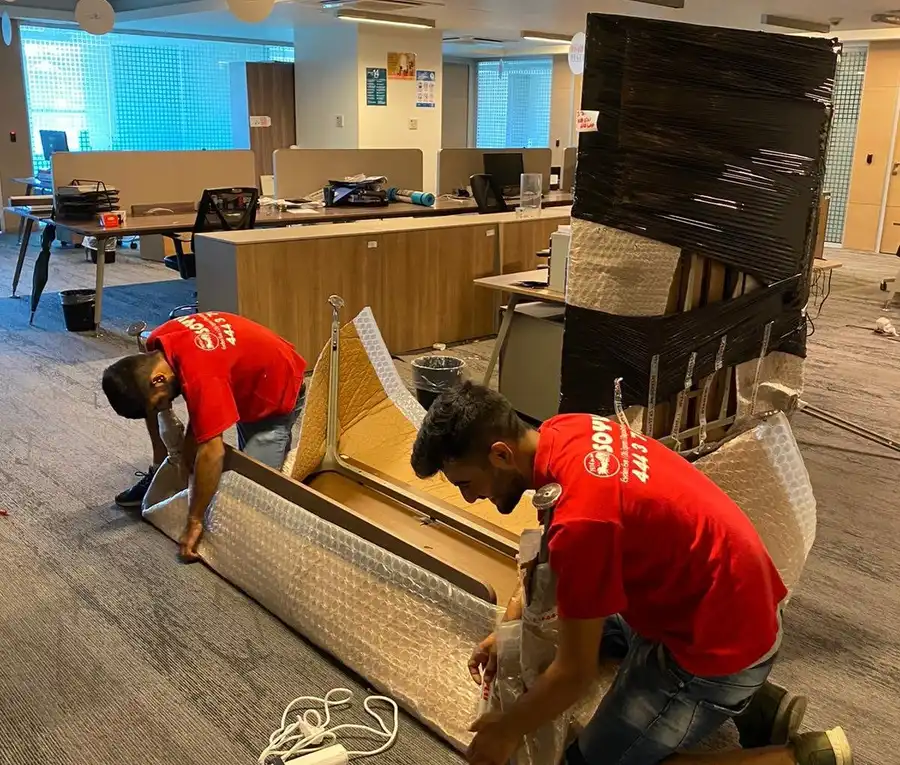 Boşaltılmakta olan açık ofiste çalışma masasını balonlu naylonla saran iki Soylu çalışanı, arkada siyah streçli dolap