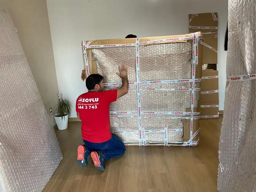 Bir Soylu çalışanı, karton köşelik ve balonlu naylonla sarılıp bantlanmış büyük, düz bir parçanın başında diz çökmüş