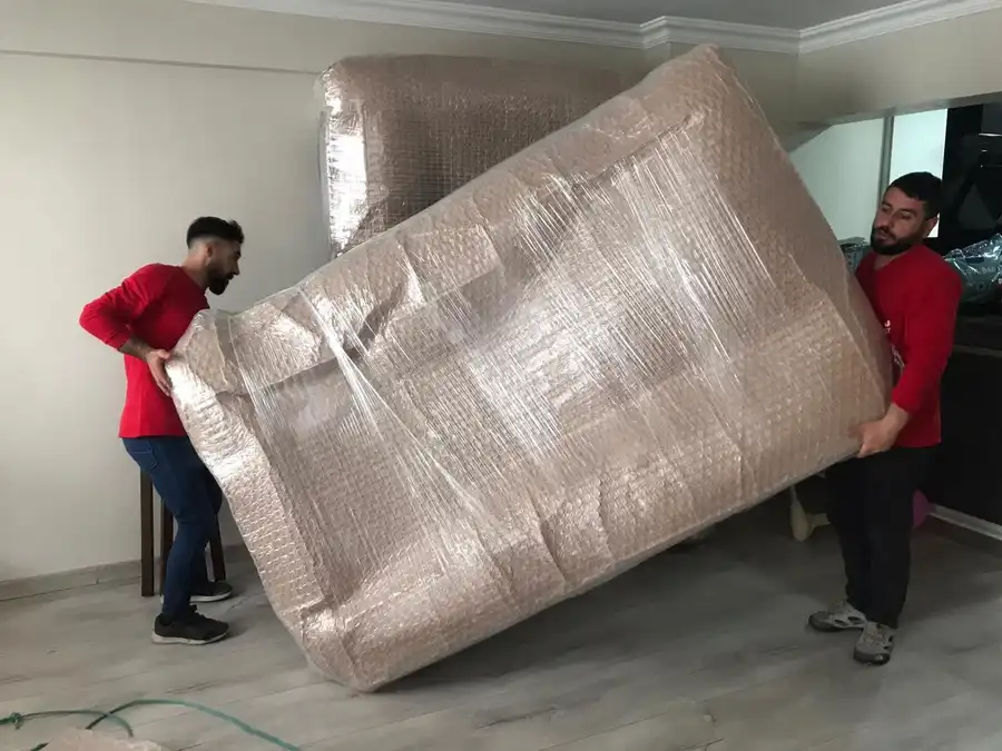 Balonlu naylon ve streç filmle sarılmış büyük bir eşyayı iki ucundan kaldıran iki taşıma personeli