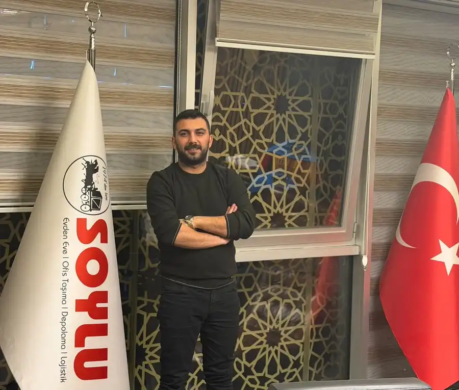 Soylu bayrağı ile Türk bayrağı arasında kollarını kavuşturmuş duran bir kişi, arkasında desenli cam ve stor perdeler