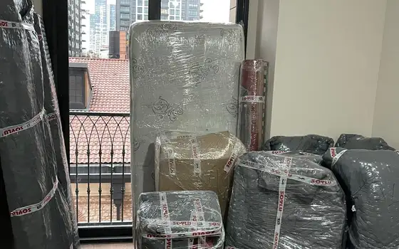 Gri battaniye ve Soylu bantlı streçle sarılmış mobilyalar, yatak ve rulo halı balkon kapısının önünde bekliyor