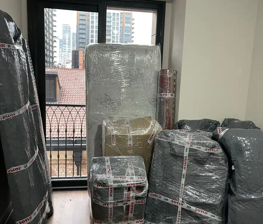 Bir odada balkon kapısının önünde streçlenmiş yatak, rulo halı ve battaniyeyle sarılmış mobilyalar