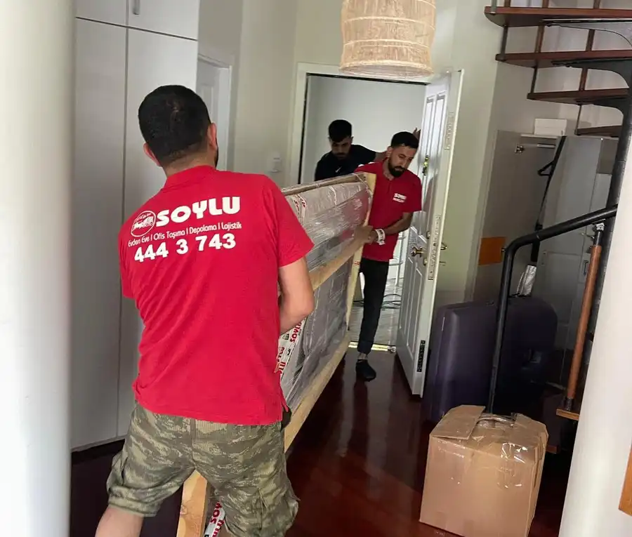 Ahşap çerçeveyle korunmuş, streç filmle sarılı uzun bir eşyayı daire kapısından içeri taşıyan kırmızı tişörtlü ekip