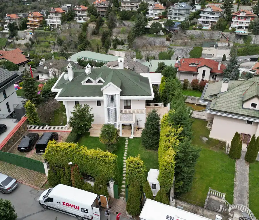 Bahçeli bir villanın önünde kapısı açık Soylu panelvanı ve kamyonu, yolda bekleyen iki kişi; havadan bakış